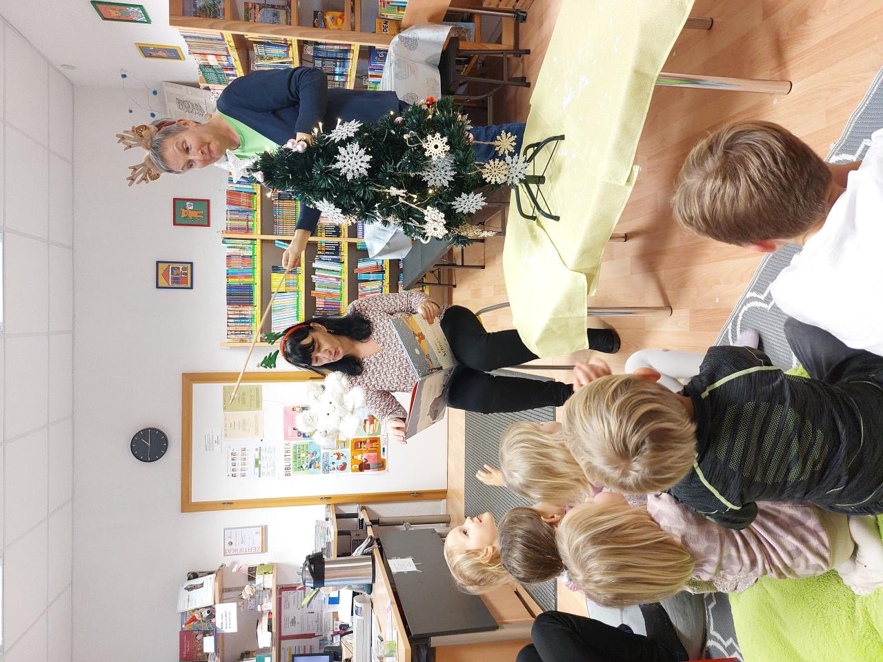 Zwei Frauen lesen Kindern etwas vor. Im Vordergrund steht ein Christbaum