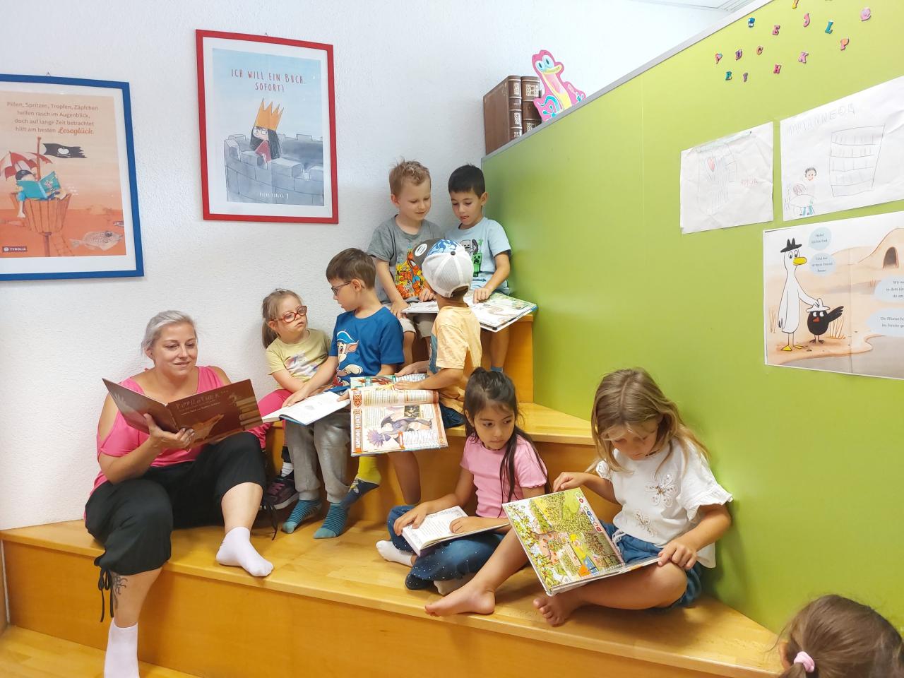 Kinder und eine Frau sitzen mit Bilderbüchern lesend auf einer Sitztreppe